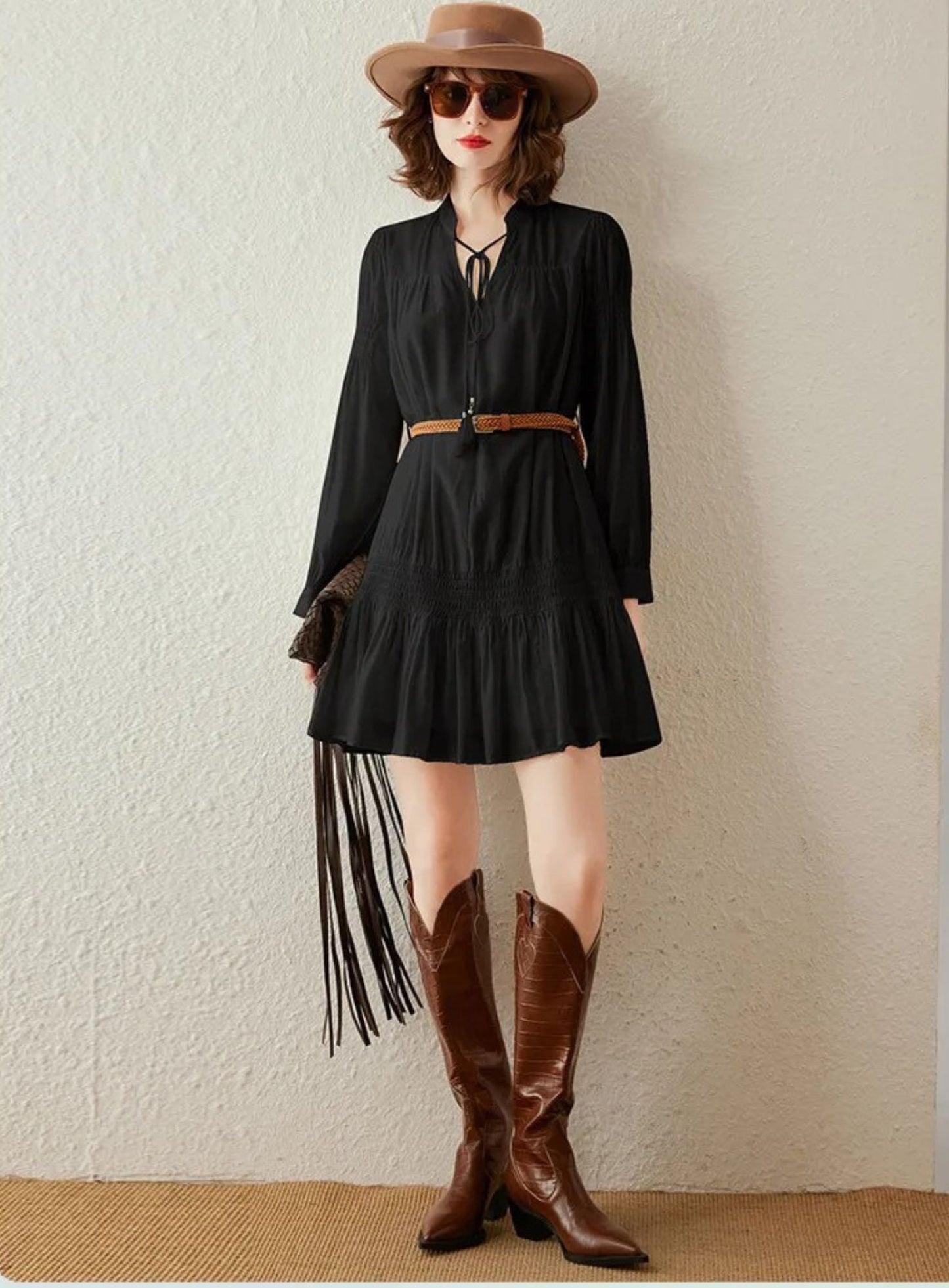 Woman wearing black long-sleeve pleated mini dress with braided brown belt, styled with hat and boots