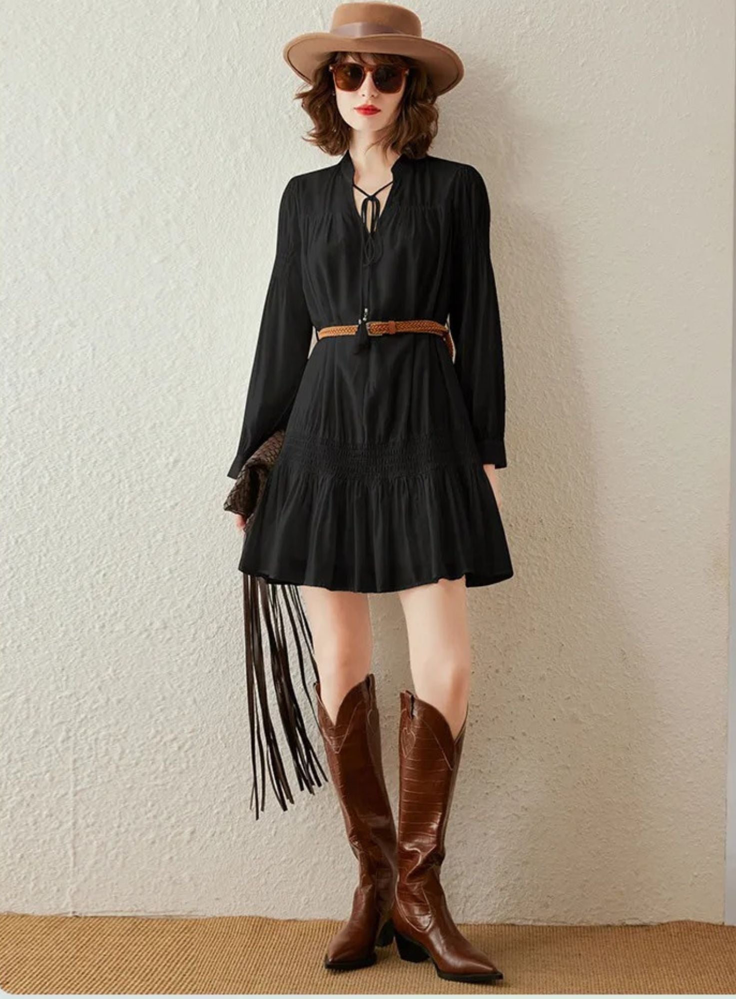Woman wearing black long-sleeve pleated mini dress with braided brown belt, styled with hat and boots