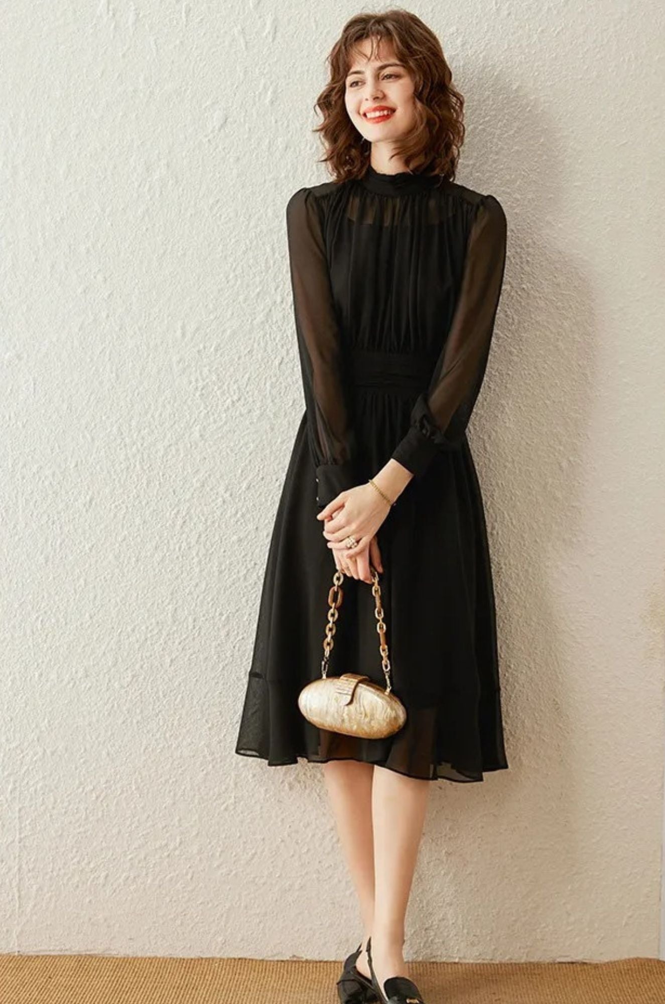 A woman wearing a black draped chiffon dress with a stand collar, sheer long sleeves, and a mid-calf A-line skirt