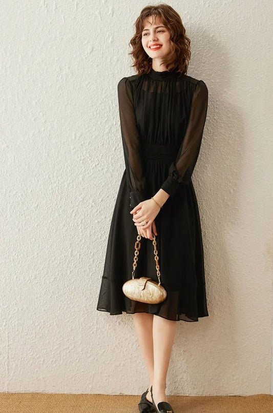 A woman wearing a black draped chiffon dress with a stand collar, sheer long sleeves, and a mid-calf A-line skirt