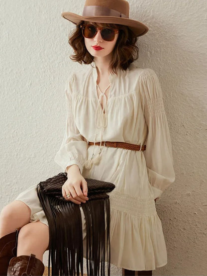 Woman wearing cream long-sleeve pleated mini dress with braided brown belt, styled with hat and sunglasses