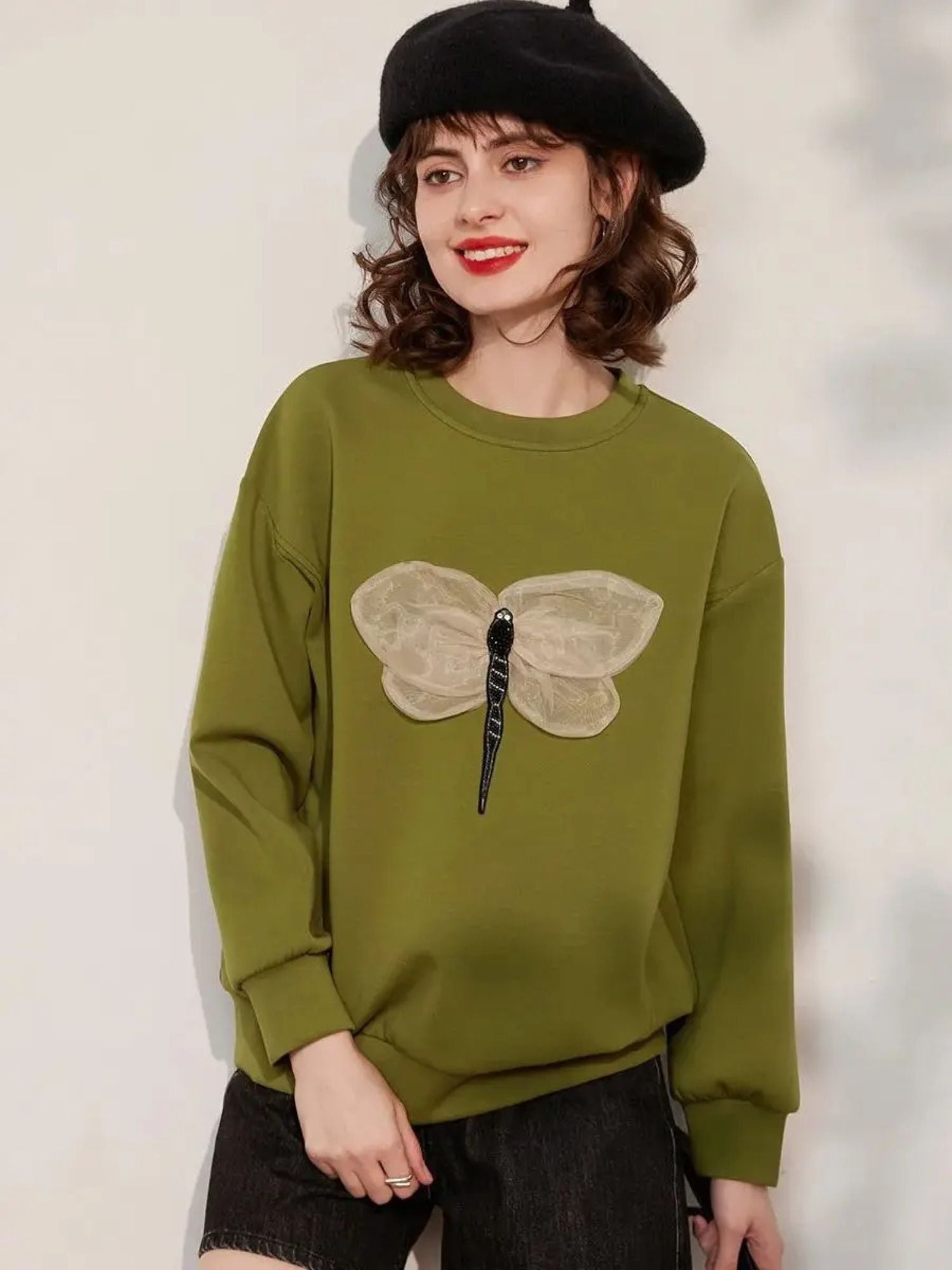 A woman wearing a green jumper with a 3D dragonfly decoration on the front, paired with black shorts