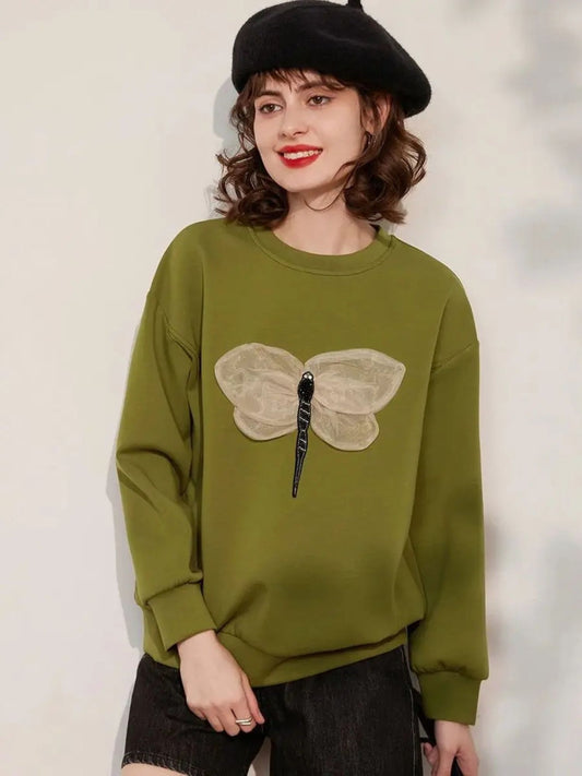 A woman wearing a green jumper with a 3D dragonfly decoration on the front, paired with black shorts