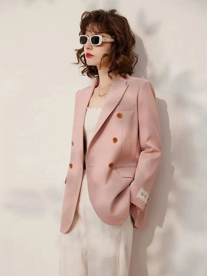 Woman wearing a blush pink double-breasted blazer with gold buttons over a white top and white trousers, accessorized with retro white sunglasses, standing in front of a neutral backdrop