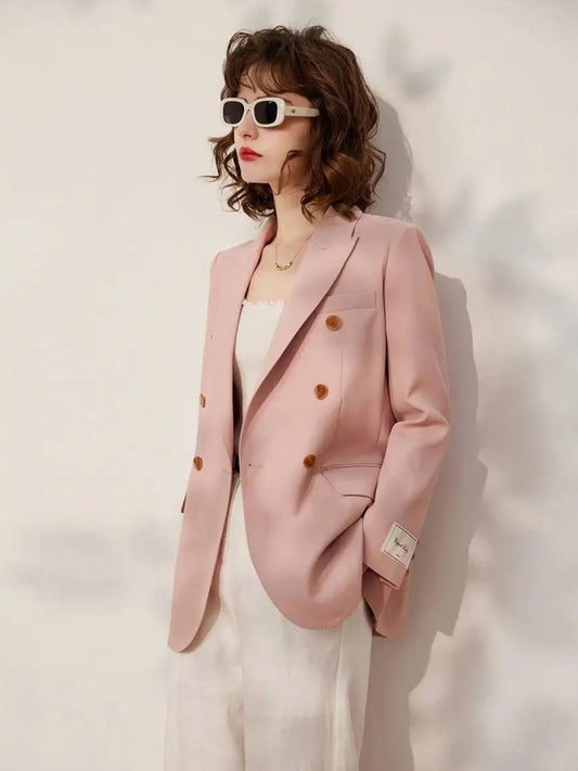 Woman wearing a blush pink double-breasted blazer with gold buttons over a white top and white trousers, accessorized with retro white sunglasses, standing in front of a neutral backdrop