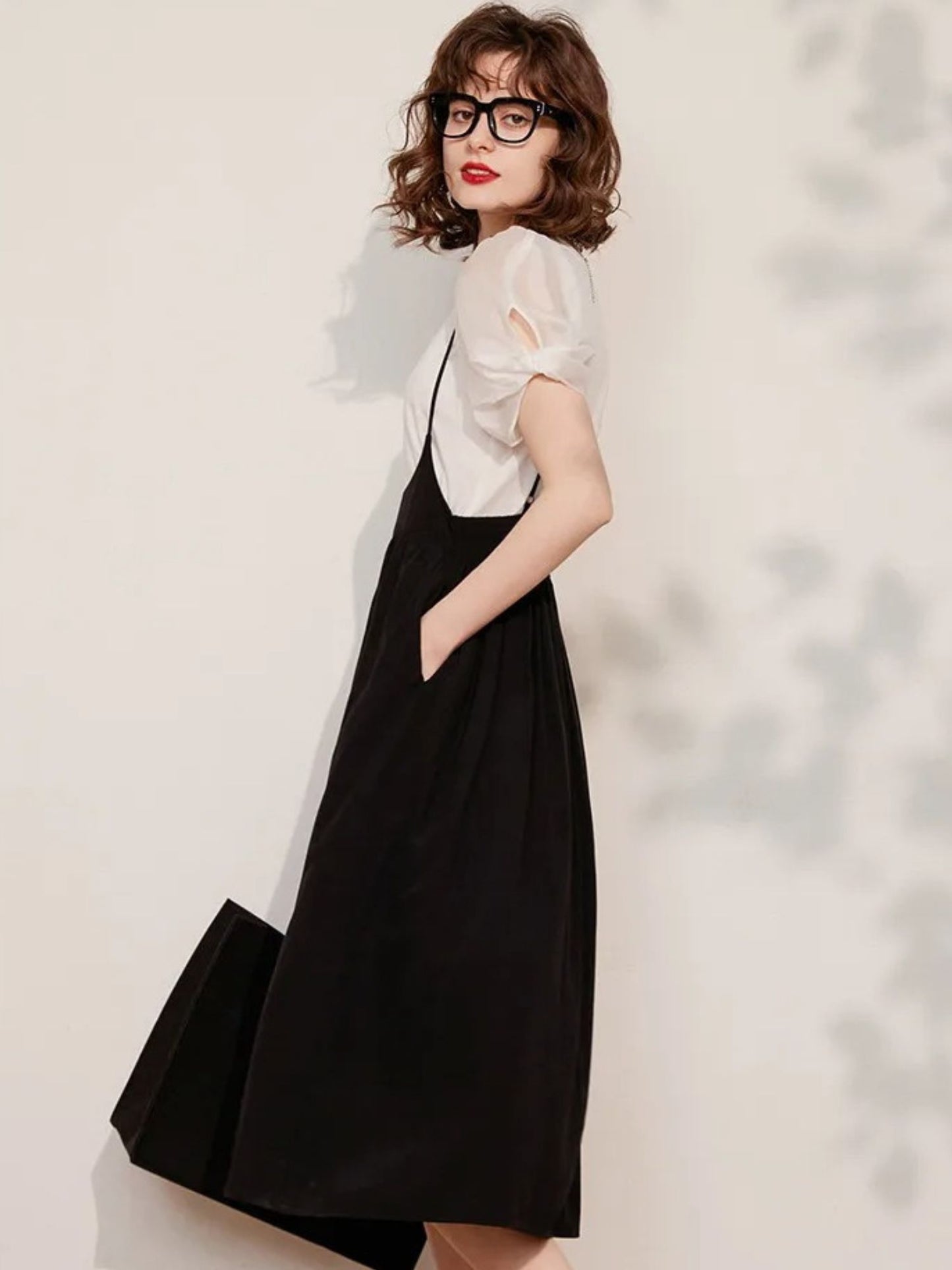 A woman wearing a black sleeveless suspender dress with an A-line silhouette and elastic waist, paired with a puff-sleeve blouse with a classic O-neckline