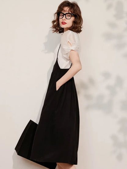 A woman wearing a black sleeveless suspender dress with an A-line silhouette and elastic waist, paired with a puff-sleeve blouse with a classic O-neckline