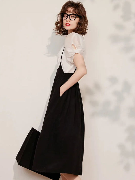 A woman wearing a black sleeveless suspender dress with an A-line silhouette and elastic waist, paired with a puff-sleeve blouse with a classic O-neckline