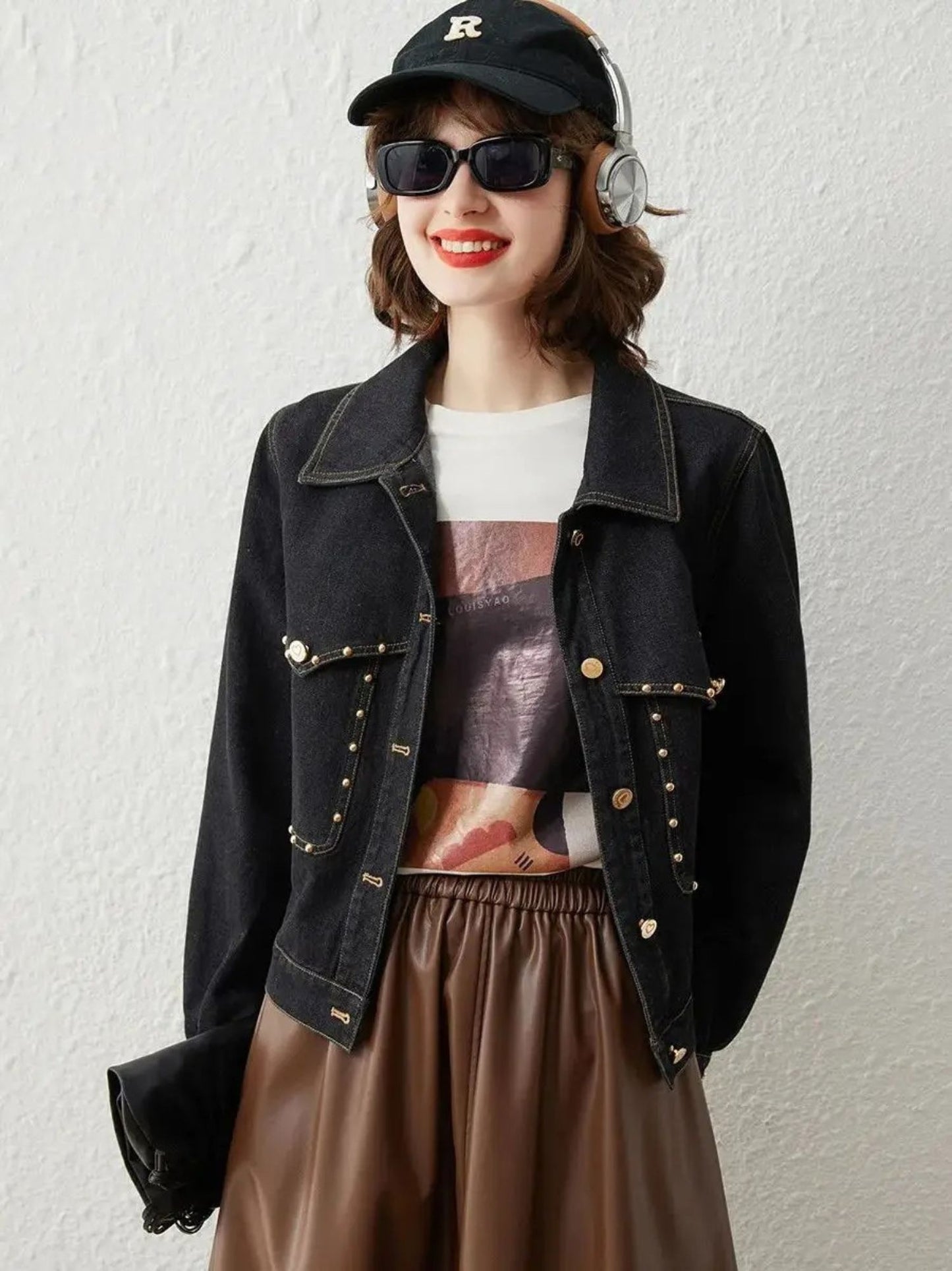 Woman wearing a black denim jacket with gold button details, sunglasses, and a cap, paired with a printed top and brown skirt