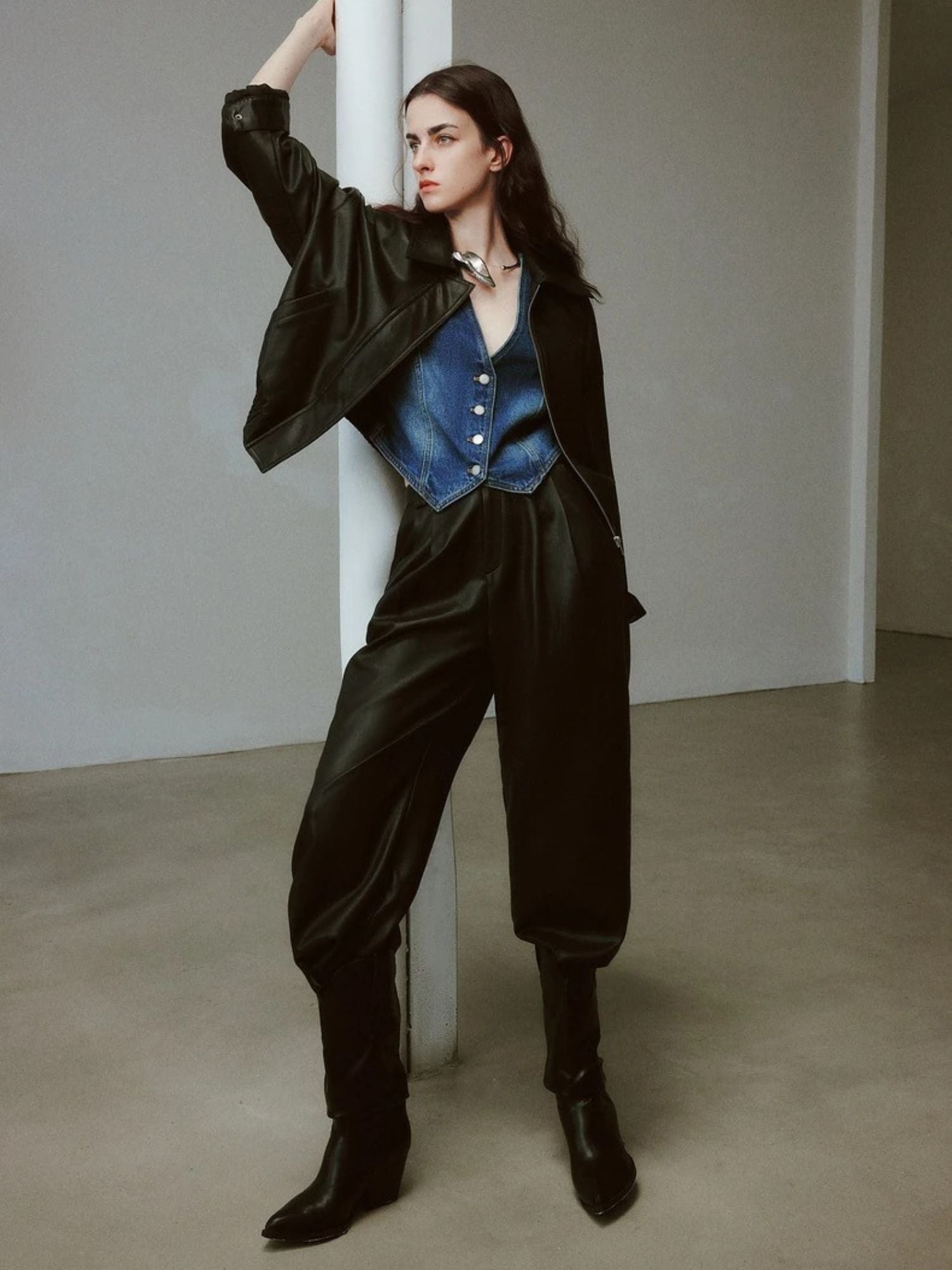 Woman wearing high-waisted black faux leather pants, blue satin vest, black jacket, and boots, posing with one arm raised