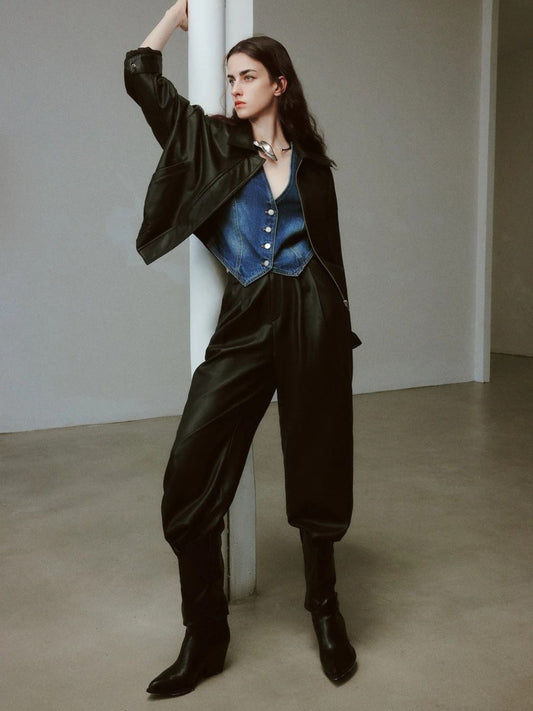 Woman wearing high-waisted black faux leather pants, blue satin vest, black jacket, and boots, posing with one arm raised