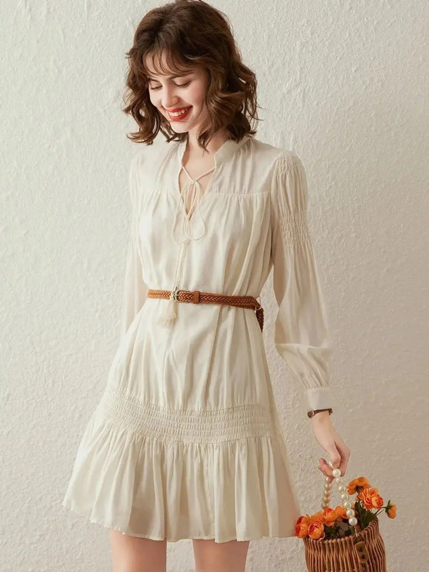 Woman in cream pleated mini dress with long sleeves, holding a brown fringe bag
