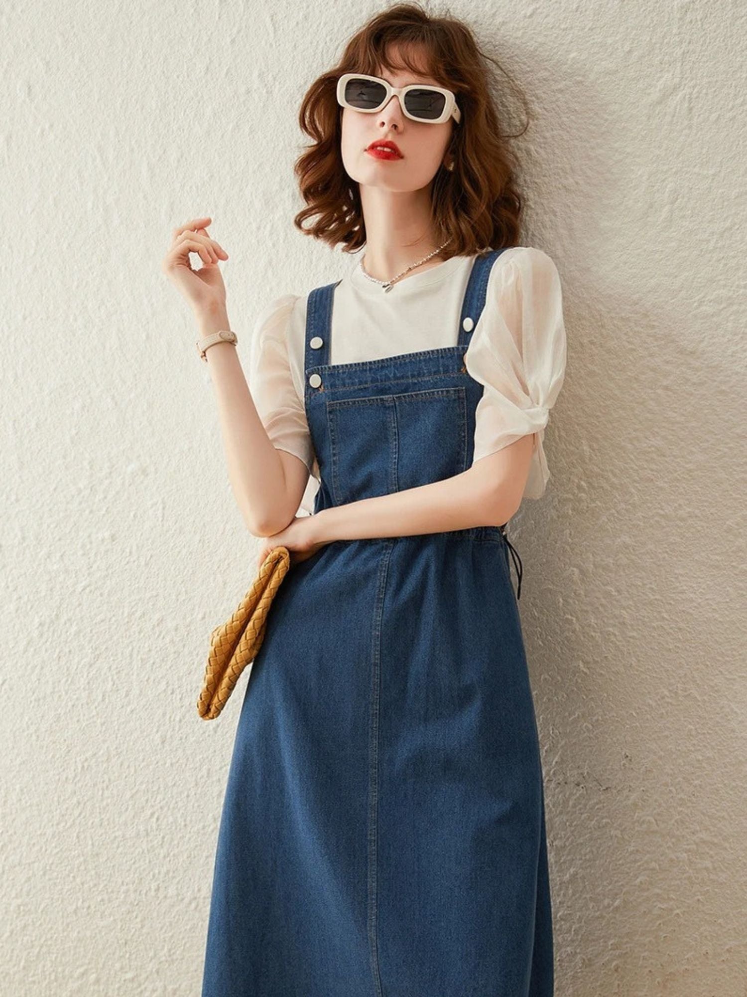 Woman wearing cream puff-sleeve blouse with denim overall dress, holding yellow clutch