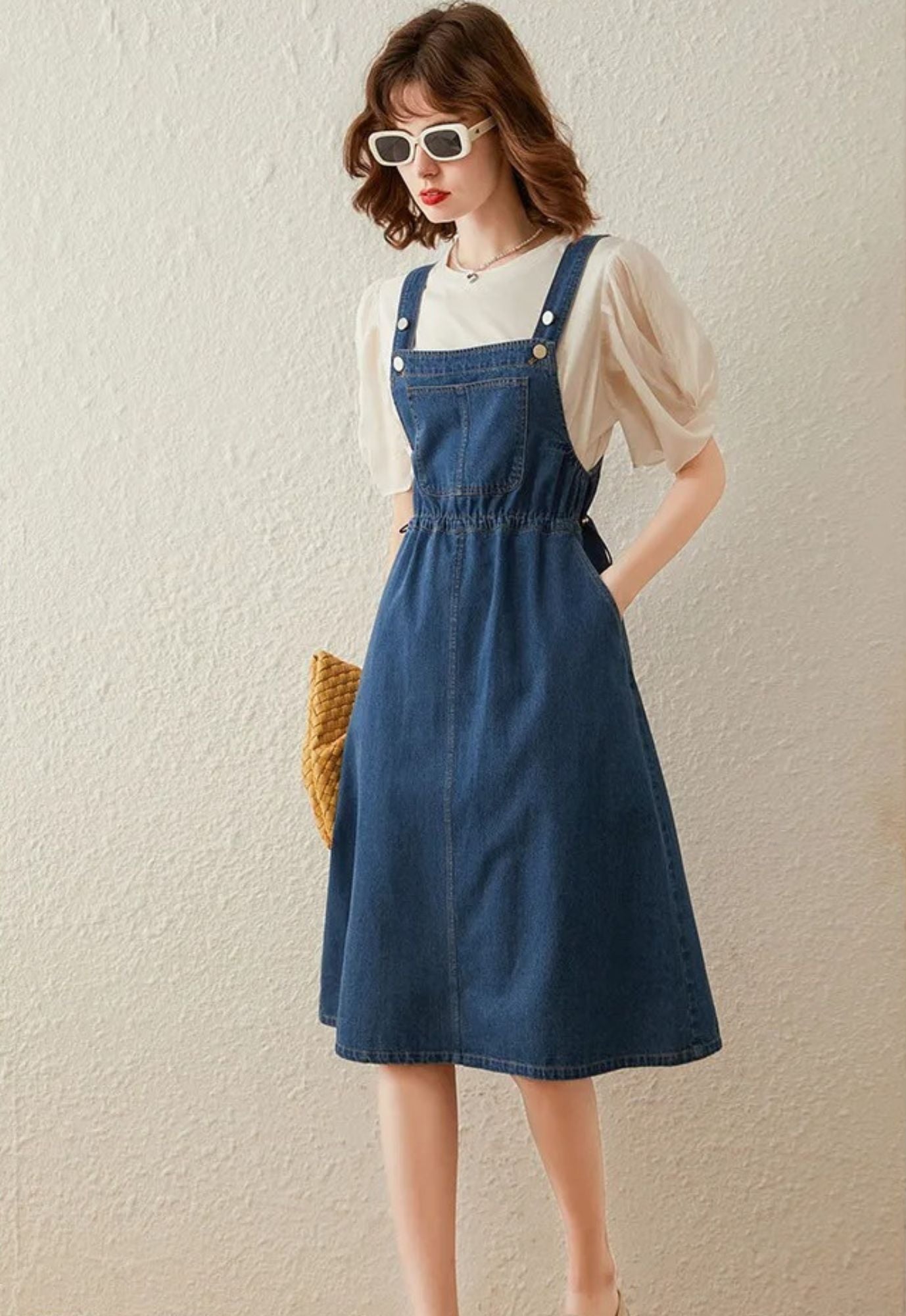 A woman wearing a blue denim suspender dress with adjustable straps, button details, and a tie at the waist