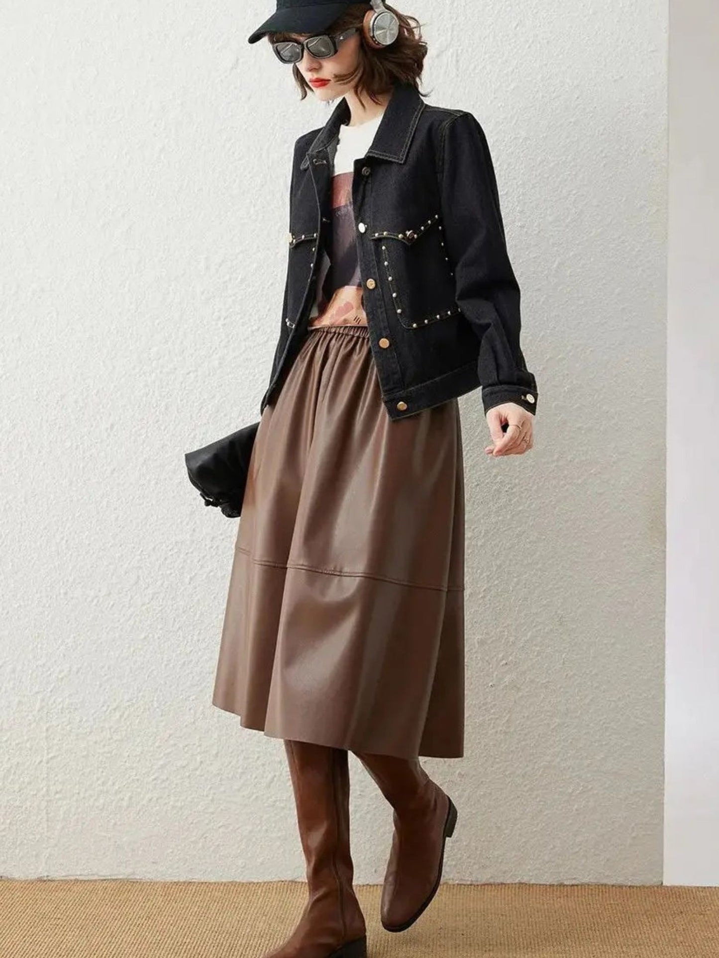 Full-body view of woman in black denim jacket, brown skirt, and tall boots, holding a bag