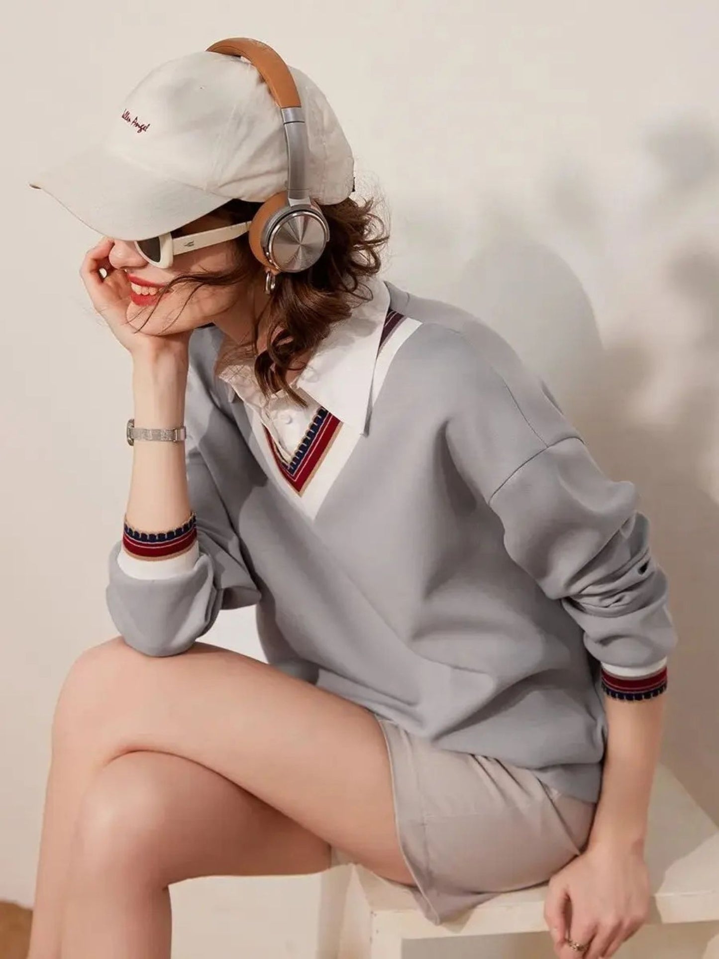 Woman seated in light grey striped collar sweatshirt and shorts, posing with cap