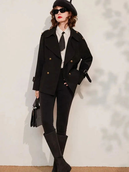 Woman leaning against wall wearing black blazer with gold buttons, white shirt, tie, and black trousers