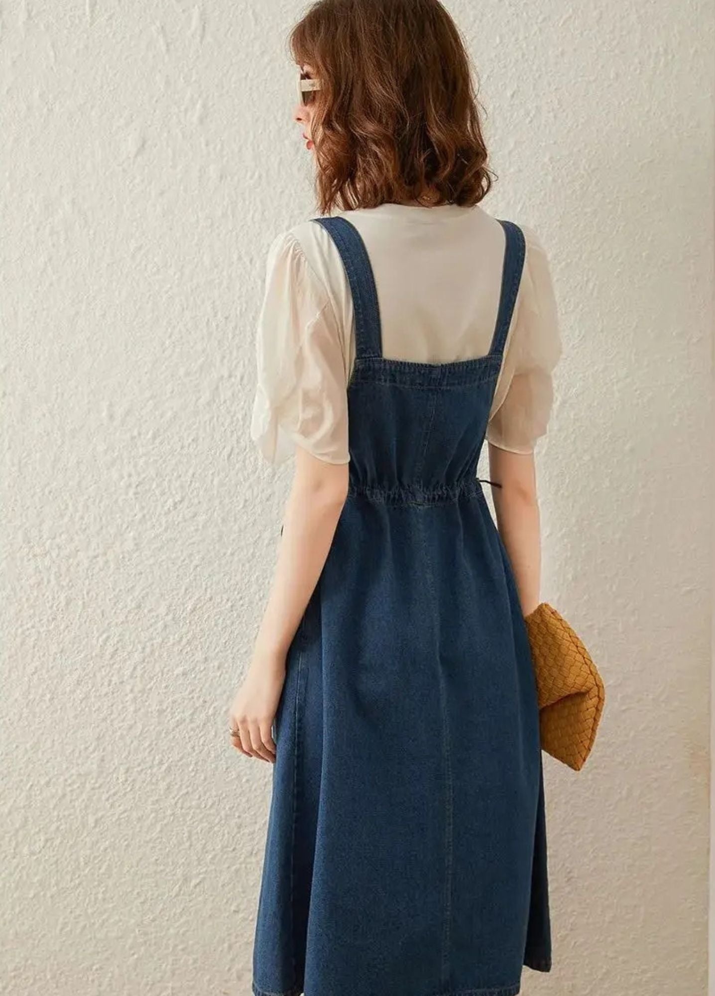Back view of a woman wearing a blue denim suspender dress with adjustable straps, button details, and a tie at the waist