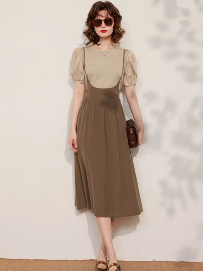 A woman wearing a khaki sleeveless suspender dress with an A-line silhouette and elastic waist, paired with a puff-sleeve blouse with a classic O-neckline