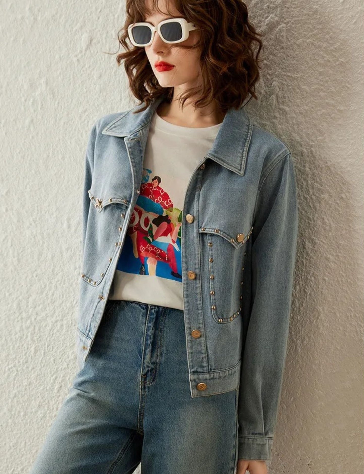 Woman in light blue denim jacket, jeans, and printed t-shirt, smiling against plain background